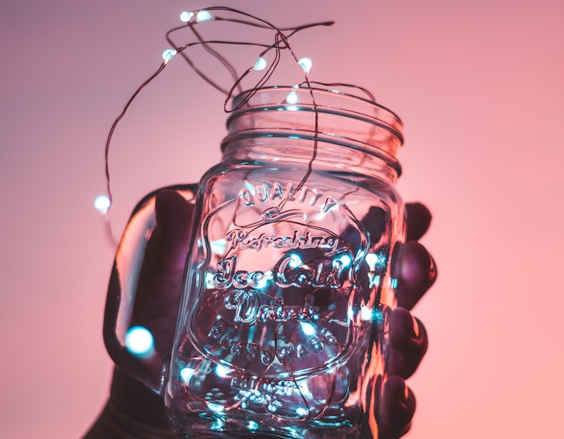 String lights in a mason jar, held by a hand against a pale pink background. 
