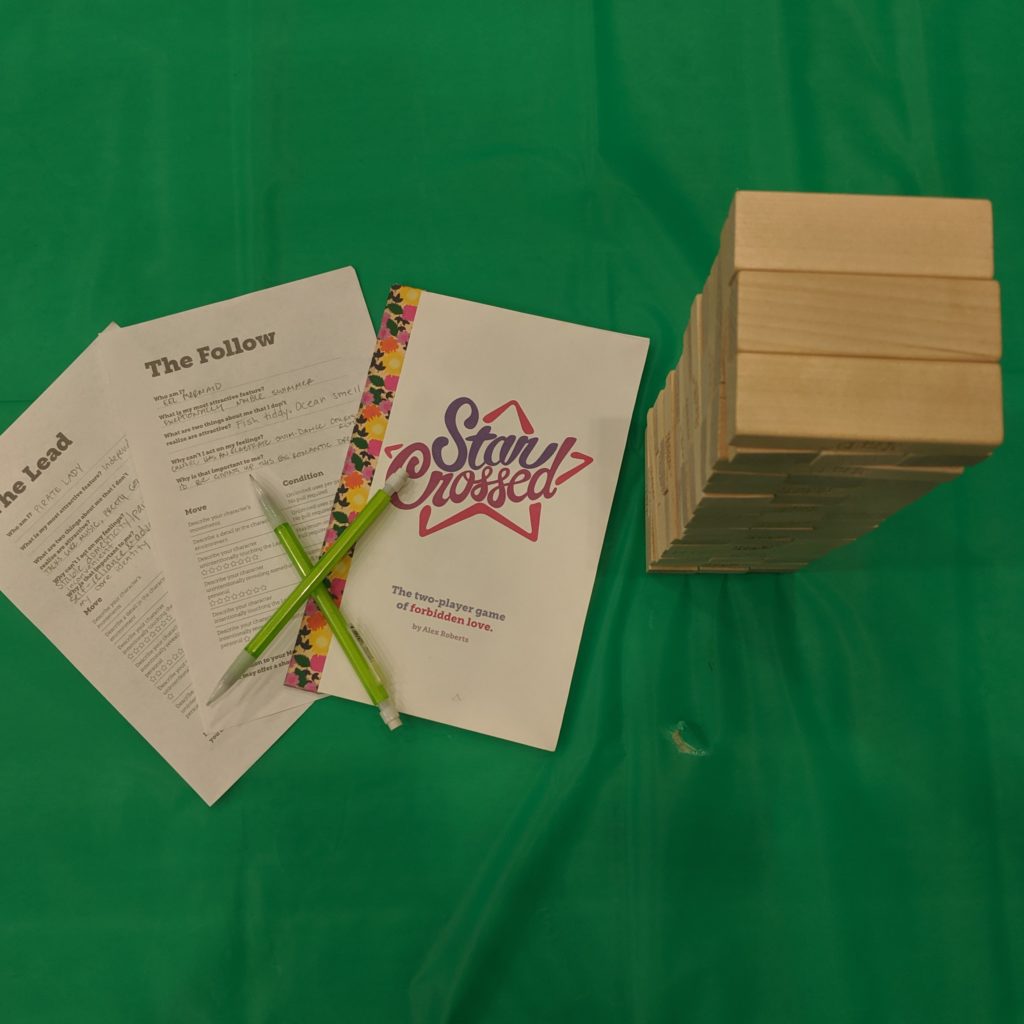 A rules pamphlet for Star Crossed, two character sheets, two pencils grossed over one another, and a Jenga tower.